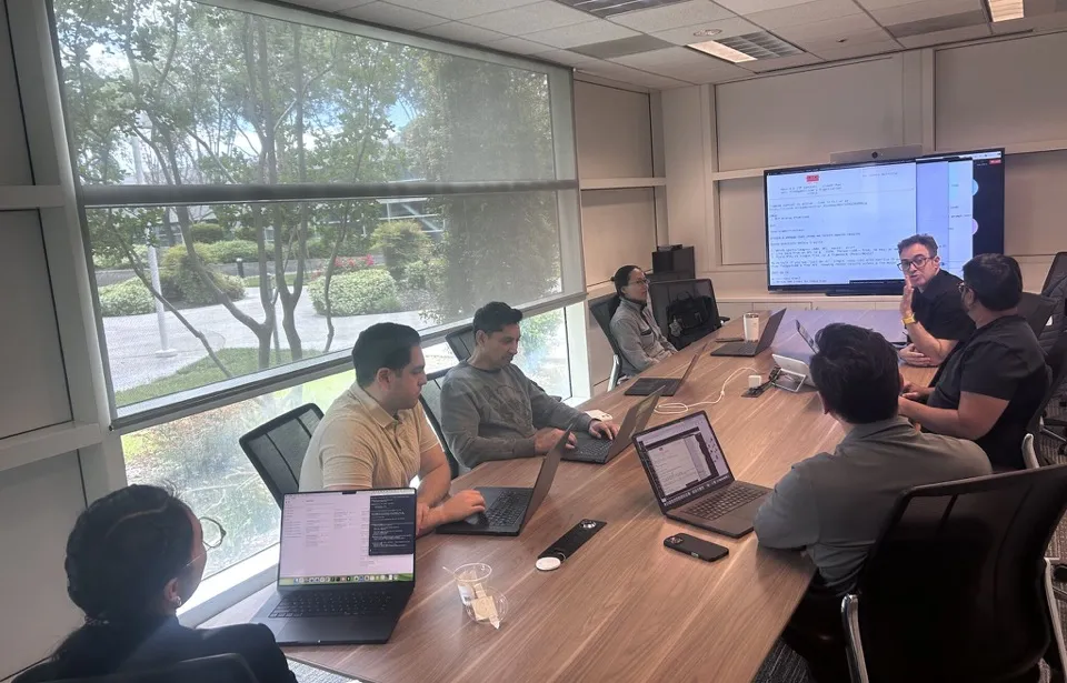A conference room at NVIDIA. Seven product managers around a long wooden table with laptops open, a wall display showing a markdown spec, Amir Fish on the right side of the table mid-conversation.